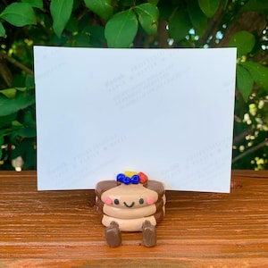 May include: A whimsical, handmade pancake figurine with a smiling face, brown pancakes, and colorful toppings. The figurine has small brown legs and is set against a white background. The overall theme is food-related and cute.