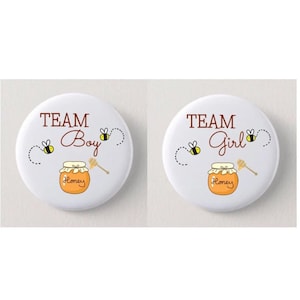 May include: Two round white buttons with black outlines. The left button says "TEAM Boy" in brown text with a honey jar and a honey dipper. The right button says "TEAM Girl" in brown text with a honey jar and a honey dipper. Both buttons have two bees flying around the text.