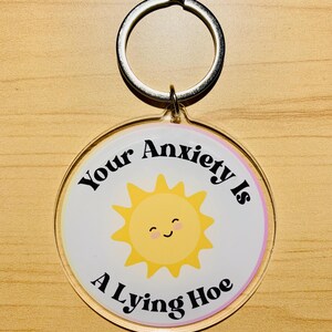 May include: A round keychain with a silver ring. The keychain has a white background with a yellow smiling sun graphic. The text "Your Anxiety Is A Lying Hoe" is printed on the keychain.