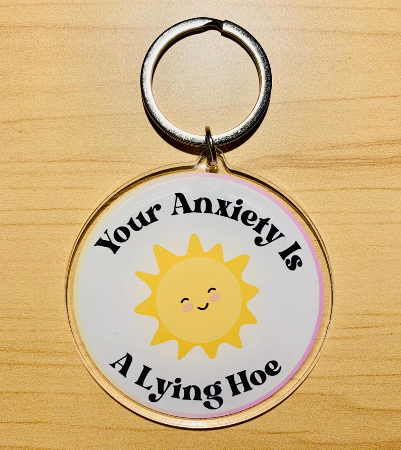 May include: A round keychain with a silver ring. The keychain has a white background with a yellow smiling sun graphic. The text "Your Anxiety Is A Lying Hoe" is printed on the keychain.
