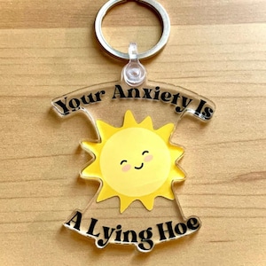 May include: A clear acrylic keychain with a silver keyring. The keychain features a yellow sun with a smiling face and the text "Your Anxiety Is A Lying Hoe" in black lettering. The keychain is on a wooden surface.