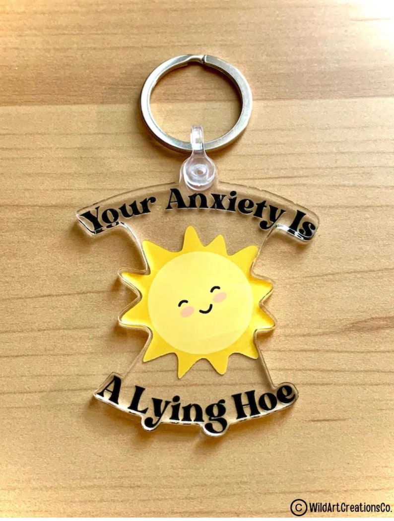 May include: A clear acrylic keychain with a silver keyring. The keychain features a yellow sun with a smiling face and the text "Your Anxiety Is A Lying Hoe" in black lettering. The keychain is on a wooden surface.