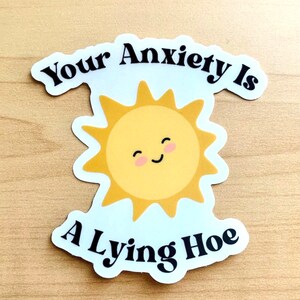 May include: A die-cut sticker featuring a smiling yellow sun with pink cheeks. The sticker has the text "Your Anxiety Is A Lying Hoe" in black, curved around the sun. The sticker is on a light wood grain surface.