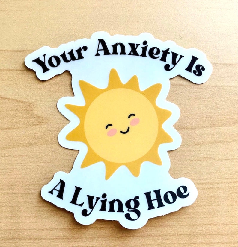 May include: A die-cut sticker featuring a smiling yellow sun with pink cheeks. The sticker has the text "Your Anxiety Is A Lying Hoe" in black, curved around the sun. The sticker is on a light wood grain surface.