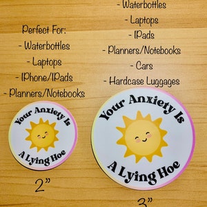 May include: Two round stickers with the text "Your Anxiety Is A Lying Hoe" and a smiling sun graphic. One sticker is 2 inches, the other is 3 inches. The stickers are white with a pink and yellow gradient border.