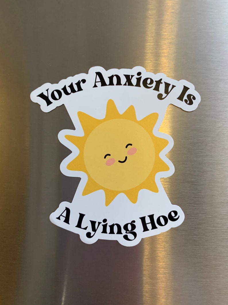 May include: A white sticker with the text "Your Anxiety Is A Lying Hoe" in black, surrounding a yellow smiling sun with a pink blush. The sticker is on a silver surface.
