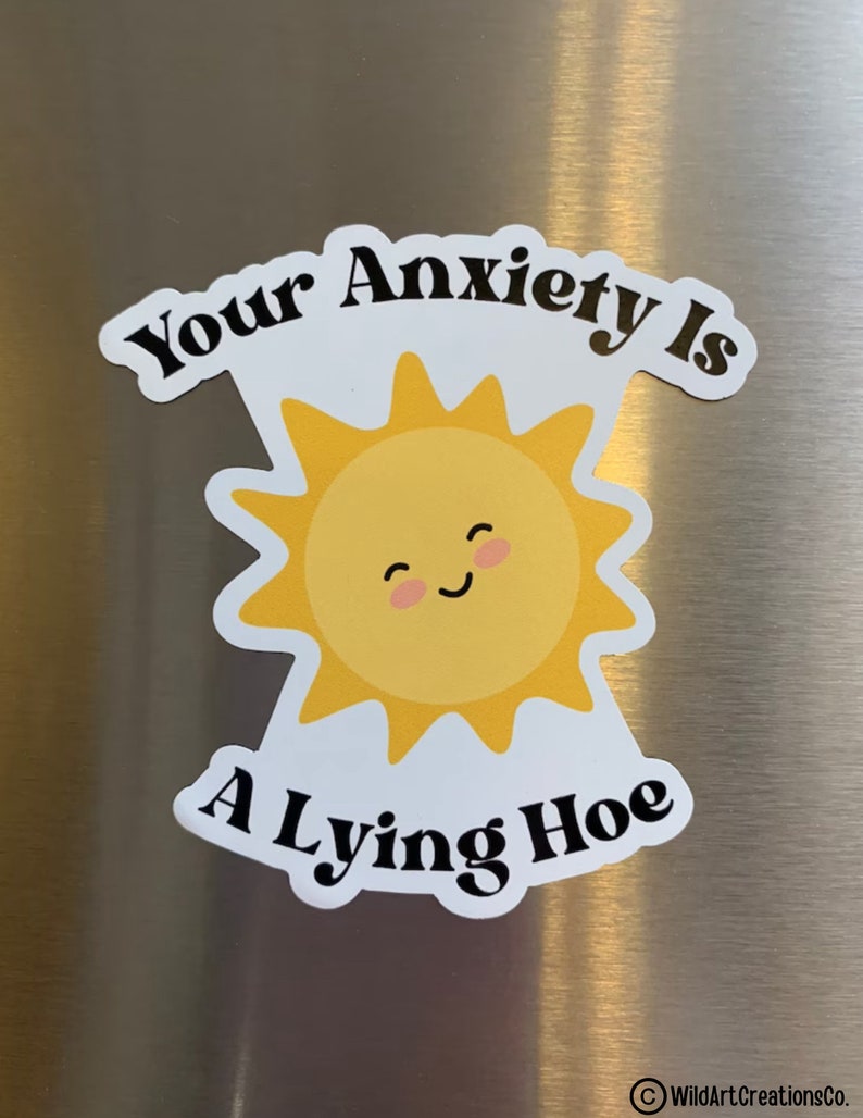 May include: A white sticker with the text "Your Anxiety Is A Lying Hoe." The sticker features a yellow sun with a smiling face. The sticker is on a metallic surface.