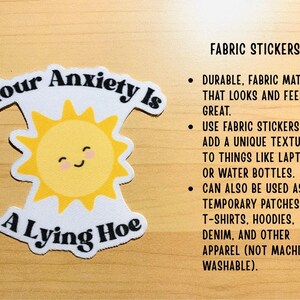 May include: A fabric sticker with the text "Your Anxiety Is A Lying Hoe" and a smiling sun graphic. The sticker is white with black text and a yellow sun. It can be used on laptops, water bottles, or apparel.