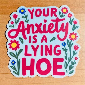 May include: A sticker with the text "YOUR Anxiety IS A LYING HOE" in red, surrounded by floral illustrations. The background is light blue, and the flowers are red, yellow, and blue.