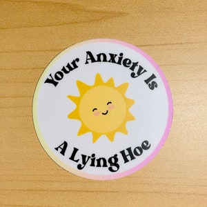 May include: A round sticker with a white background and a yellow, smiling sun graphic. The text "Your Anxiety Is A Lying Hoe" is printed in black around the sun. The sticker has a gradient pink and yellow border.