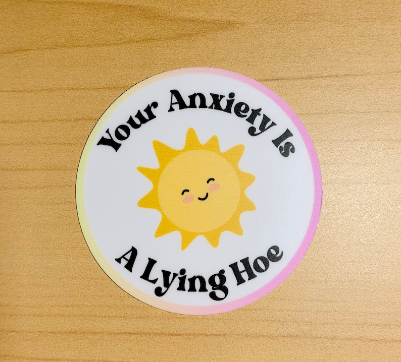 May include: A round sticker with a white background and a yellow, smiling sun graphic. The text "Your Anxiety Is A Lying Hoe" is printed in black around the sun. The sticker has a gradient pink and yellow border.