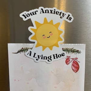 May include: A white magnet with the text "Your Anxiety Is A Lying Hoe" and a smiling yellow sun graphic. The magnet is placed over a watercolor-style paper with festive decorations.