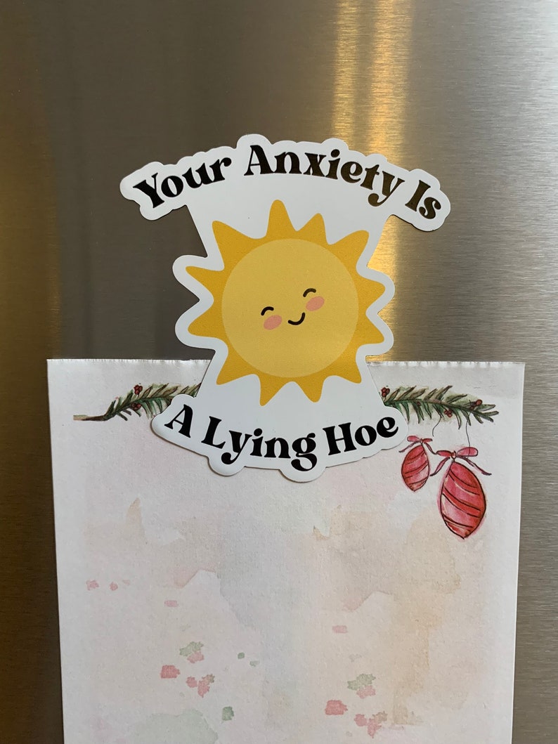 May include: A white magnet with the text "Your Anxiety Is A Lying Hoe" and a smiling yellow sun graphic. The magnet is placed over a watercolor-style paper with festive decorations.