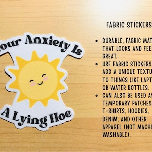 May include: A fabric sticker with the text "Your Anxiety Is A Lying Hoe" and a smiling sun graphic. The sticker is white with black text and a yellow sun. It can be used on laptops, water bottles, or apparel.