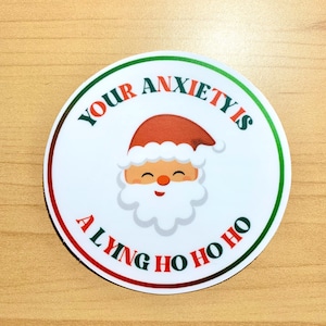 May include: A round sticker with a Santa Claus graphic and the text "Your Anxiety Is A Lying Ho Ho Ho". The Santa has a red hat, a white beard, and a smiling face. The text is in red and green.