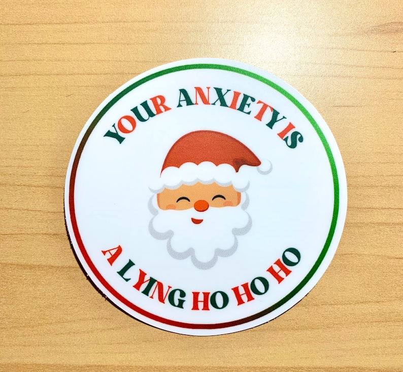 May include: A round sticker with a Santa Claus graphic and the text "Your Anxiety Is A Lying Ho Ho Ho". The Santa has a red hat, a white beard, and a smiling face. The text is in red and green.