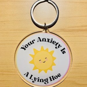 May include: A round keychain with a silver ring. The keychain has a white background with a yellow smiling sun graphic. The text "Your Anxiety Is A Lying Hoe" is printed around the sun in black.