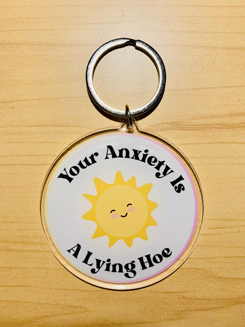 May include: A round keychain with a silver ring. The keychain has a white background with a yellow smiling sun graphic. The text "Your Anxiety Is A Lying Hoe" is printed around the sun in black.