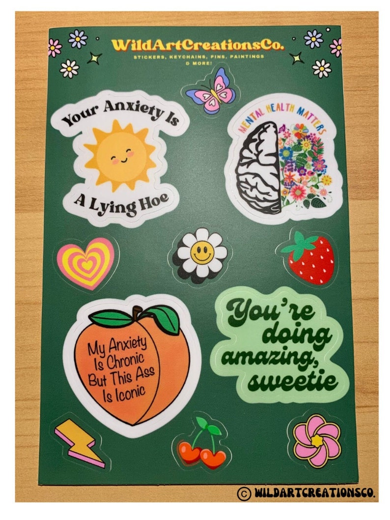 May include: A sheet of stickers with various designs and text. Designs include a smiling sun, a brain with flowers, a peach with text, and a daisy. Text includes "Your Anxiety Is A Lying Hoe" and "Mental Health Matters".