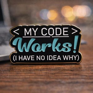 May include: A black and gold enamel pin with the text "MY CODE Works!" in white and teal. Below the text is "(I HAVE NO IDEA WHY)". The pin is on a wooden surface.