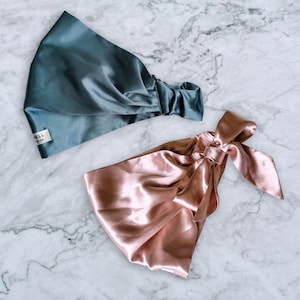 May include: Two satin hair wraps in teal and rose gold. The teal wrap features a gathered elastic band and a small white tag with text. The rose gold wrap includes a decorative tied bow detail on one side, showcasing a smooth, reflective finish.