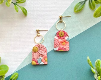 May include: A pair of floral-themed earrings. Each earring features a gold-colored post and a circular accent. The main body of each earring is a pink arch shape with embossed flowers in shades of pink, yellow, and blue. The earrings are set against a backdrop of white, teal, and blue.