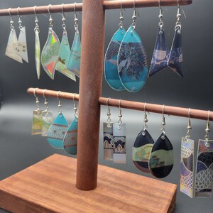 Puede incluir: Un expositor de pendientes de madera con una variedad de pendientes que presentan diferentes patrones y colores. Los pendientes están hechos de vidrio y tienen ganchos plateados.