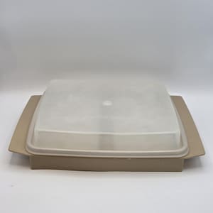 May include: A square, tan-colored food storage container with a clear, frosted lid. The container has a rectangular base with a slightly raised rim and a matching lid. The lid has a small, circular handle in the center.