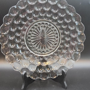 May include: Clear glass plate with a bubble design and scalloped edges. The center features a sunburst pattern. The plate is displayed on a black stand, showcasing its intricate details and texture. A decorative piece for serving or display.