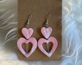 Peut inclure: Une paire de boucles d'oreilles en forme de cœur rose avec un motif de tourbillon blanc. Les boucles d'oreilles sont en argile polymère et ont des crochets argentés.