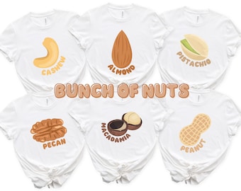 May include: Six white t-shirts featuring illustrations of nuts including a cashew, almond, pistachio, pecan, macadamia, and peanut. Each shirt displays the name of the nut in brown text below the graphic. The center text reads BUNCH OF NUTS.