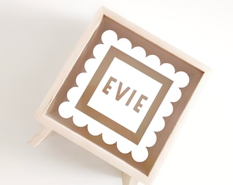 Puede incluir: Una caja de madera blanca con un frente de vidrio transparente y un marco blanco festoneado. La caja tiene un fondo marrón con el nombre "EVIE" escrito en letras blancas.
