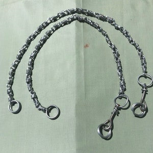 May include: Two silver-colored metal chain handles with circular end loops. The chains are made of small, tightly wound wire links. The handles are likely for a bag or purse.