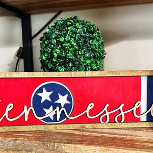 May include: A wooden sign with a red, white, and blue Tennessee state flag design. The word "Tennessee" is written in gold cursive lettering on the sign.