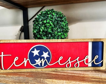 May include: A wooden sign with a red, white, and blue Tennessee state flag design. The word "Tennessee" is written in gold cursive lettering on the sign.