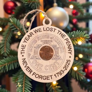 May include: A wooden Christmas ornament with a penny at the center, featuring the text "The Year We Lost The Penny" and "Never Forget 1793-2025". The ornament is hanging on a Christmas tree with red and gold ornaments.