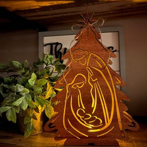 May include: A wooden Christmas tree ornament with a cutout design of the nativity scene. The ornament is illuminated from within, casting a warm glow.
