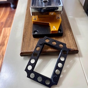 May include: A metal button maker with a silver top, gold and black base, and a black frame with circular holes. The button maker sits on a wooden base.