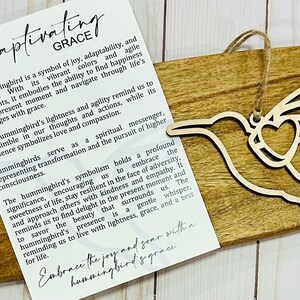 May include: Wooden hummingbird ornament with a heart cutout, hanging from a string. The ornament is on a wooden surface with a white card that reads "Captivating Grace" and includes a quote about the hummingbird's symbolism of joy, adaptability, resilience, and lightness. The quote also mentions the hummingbird's ability to find happiness in the present moment and navigate through life's challenges with grace.
