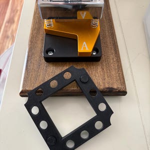 May include: A metal button maker with a silver top, gold and black base, and a wooden platform. A black square frame with circular holes is in the foreground. The letter "A" is visible on the gold base.
