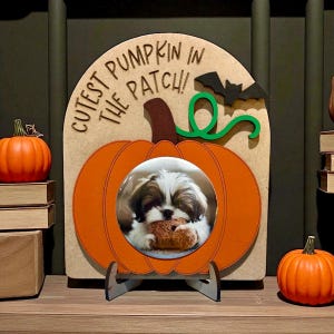 May include: A wooden pumpkin-shaped photo frame with the text "Cutest Pumpkin in the Patch!" and a small bat cutout. The frame holds a photo of a small dog. Two orange pumpkins are on either side of the frame.