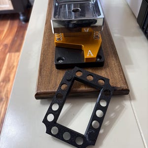 May include: A metal and gold press machine with a black frame. The machine has a silver top and is mounted on a wooden base. A black, rectangular frame with circular holes is in front of the machine.