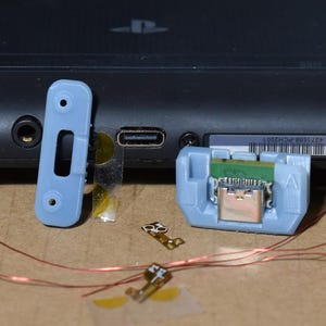 May include: A black handheld gaming console with disassembled components. A light blue plastic piece, a small circuit board, and copper wires are next to the console. The console has a PlayStation logo.