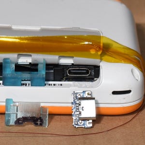 May include: Close-up of a white and orange electronic device with a section opened to reveal internal components. A blue plastic piece and a small circuit board are visible. A yellow tape secures the opened section.