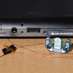 May include: A black and silver handheld gaming console, with a textured back and a series of ports and labels along the bottom edge. Several small electronic components and wires are placed near the console.