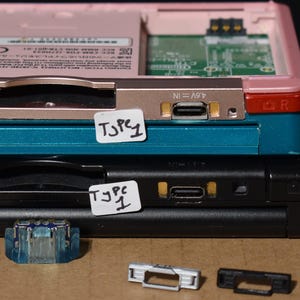 May include: Stacked Nintendo 3DS consoles in pink, teal, and black. The top console is pink and red, the middle is teal, and the bottom is black. White labels read "3DS", "3DS XL", and "Type 1".