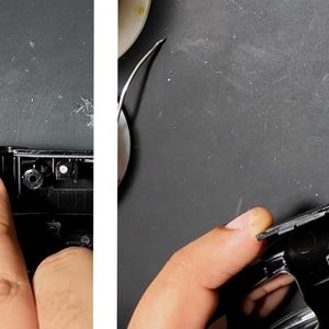 May include: Close-up of a person's finger pointing at a black plastic component. The component is part of a larger black device, possibly a game console or electronic gadget. A small black piece is detached.