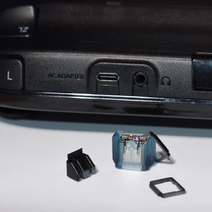 May include: Close-up of a black electronic device with the text "AC ADAPTER" and various ports. Several small, detached components are in front of the device, including a translucent blue and clear plastic piece and a black plastic piece. The device appears to be a game console.