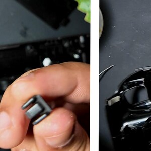 May include: Close-up of a black plastic component held in a hand, with a matching black electronic device in the background. The component has a rectangular shape with two prongs. The device has multiple small, white, circular components.