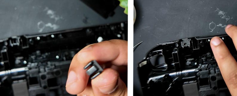 May include: Close-up of a black plastic component held in a hand, with a matching black electronic device in the background. The component has a rectangular shape with two prongs. The device has multiple small, white, circular components.